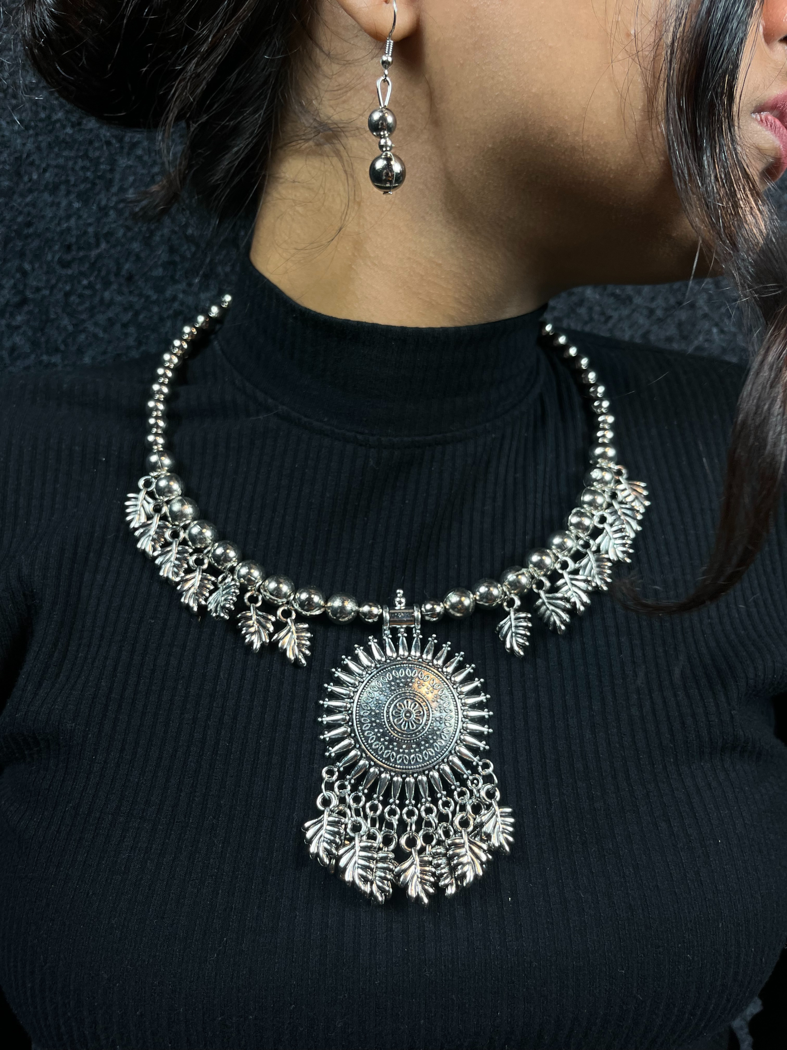 Woman wearing an oxidized silver statement necklace set featuring a large round pendant with tribal designs and matching drop earrings, perfect for ethnic and festive occasions.