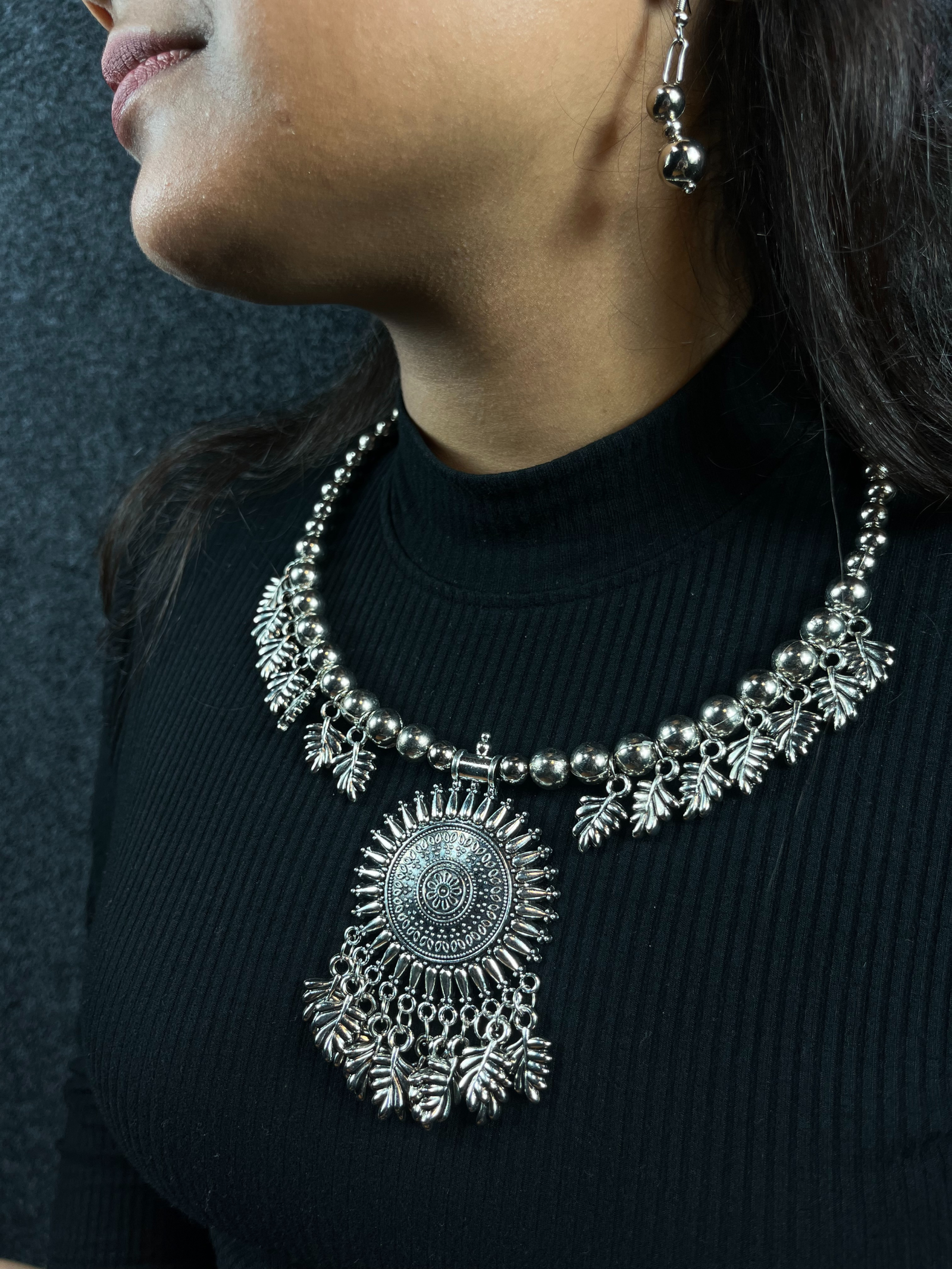 Woman wearing an oxidized silver statement necklace set featuring a large round pendant with tribal designs and matching drop earrings, perfect for ethnic and festive occasions.